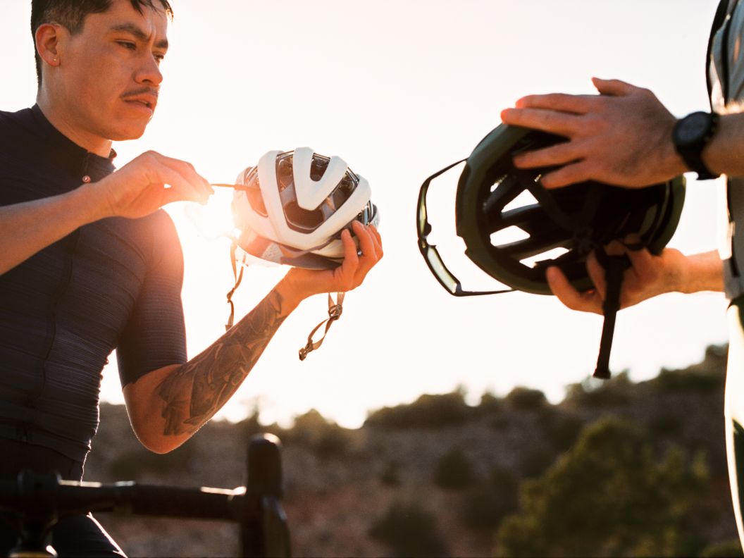Two road cyclists hold their helmets as they prepare for a ride at golden hour.