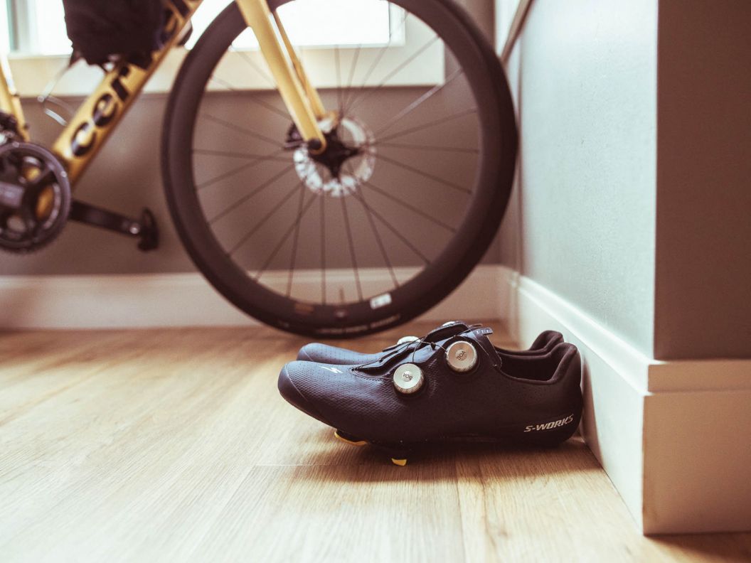 S-Works torch shoes rest together on a hardwood floor next to baseboards. A Cervelo Soloist road bike is against the wall further back.