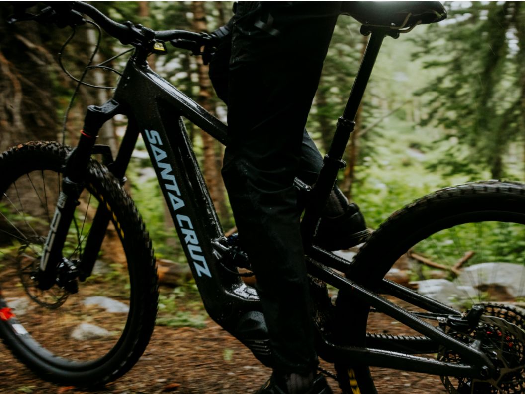 A rider on a Santa Cruz E-MTB stands over it in a forest. 