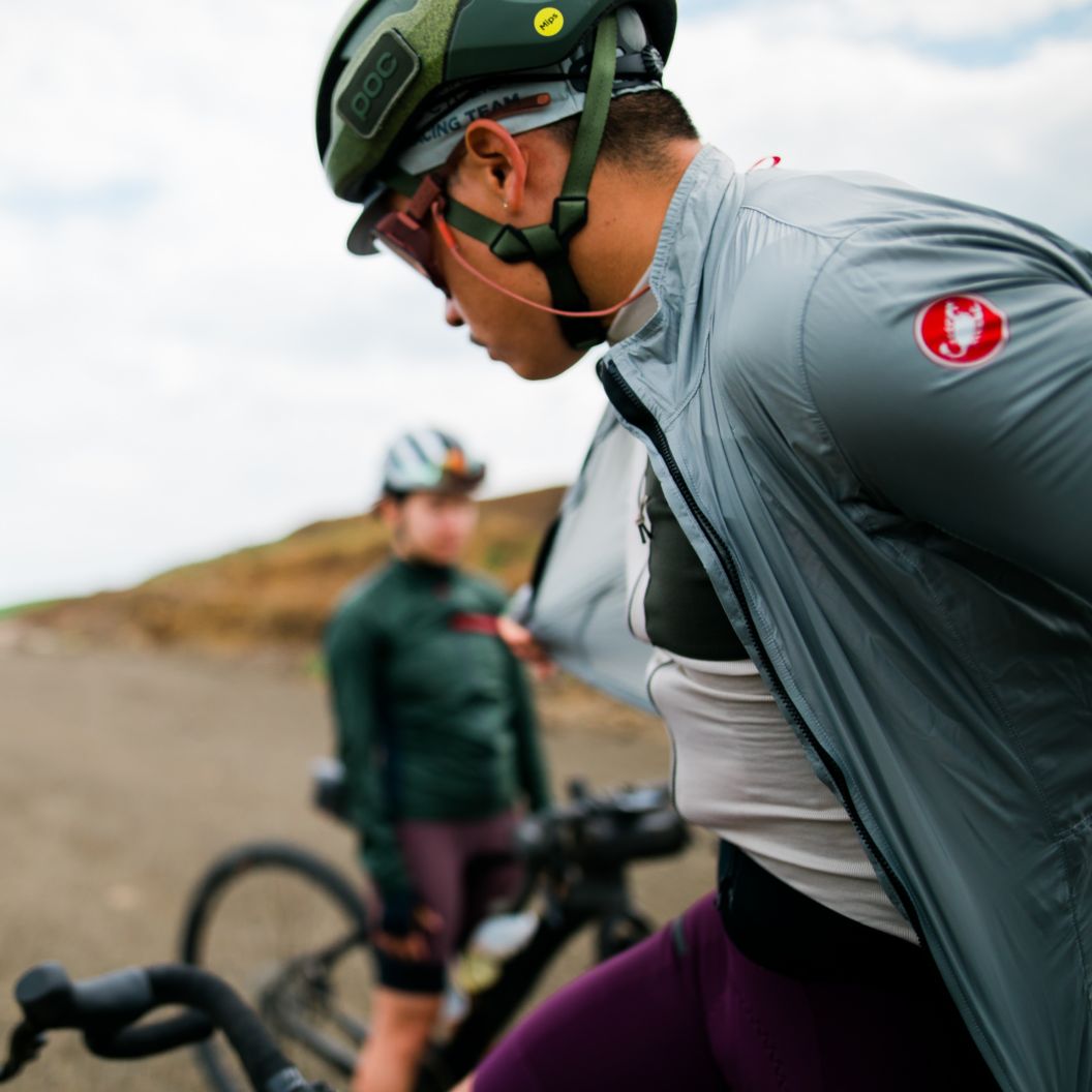 Two gravel riders take a break in an open area and one removes their Castelli wind jacket.