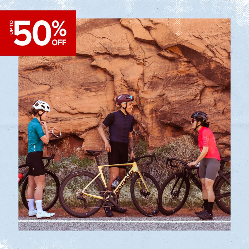 Three cyclists stand with their bikes and converse next to a red wall of rock. 