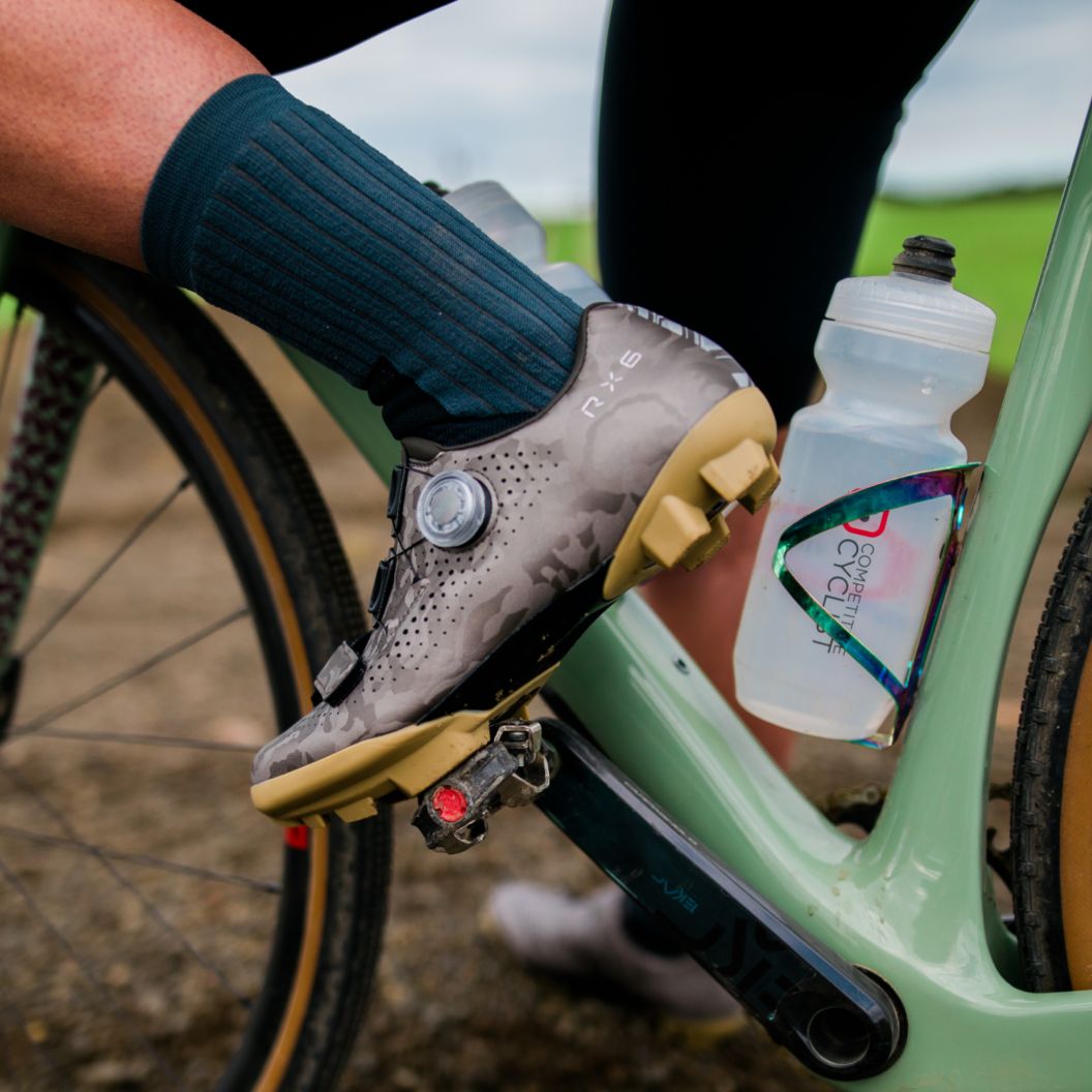 A rider in gravel shoes stands over a green bike with one foot clipped into the pedal.