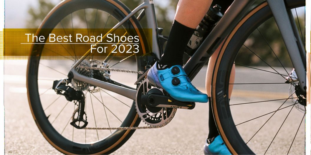 A cyclist in blue Shimano shoes has their right foot clipped into the pedals while standing over their bike.