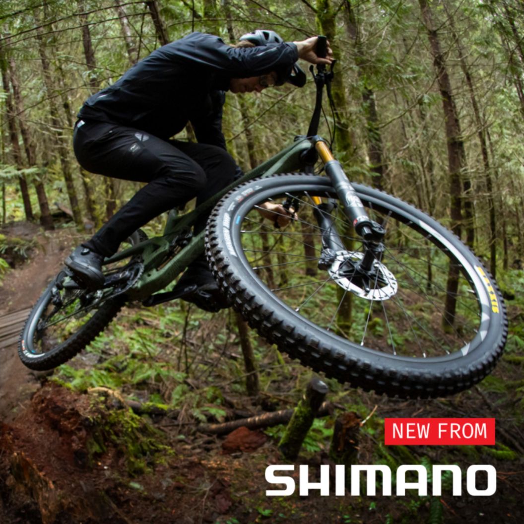 A mountain biker soars over a berm on a lush forest trail. The superimposed text reads: New From Shimano.