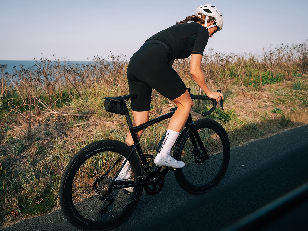 Two cyclists riding a coastal road with white chalk cliffs and dry grassland in the background, ocean visible in the distance