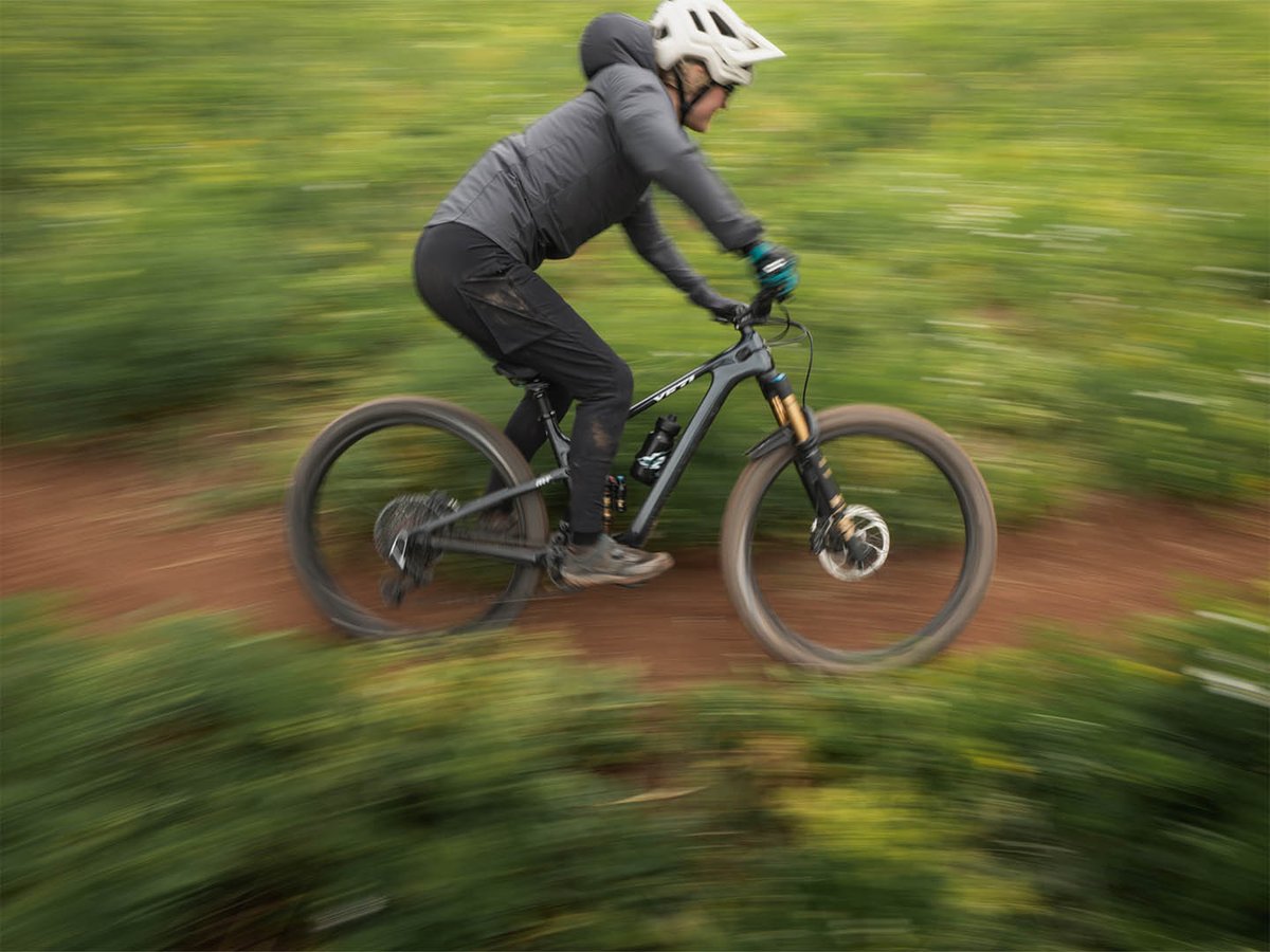 Panning shot of a mountain biker riding fast on a carbon Yeti bike, green brush blurred on both sides of the dirt trail.