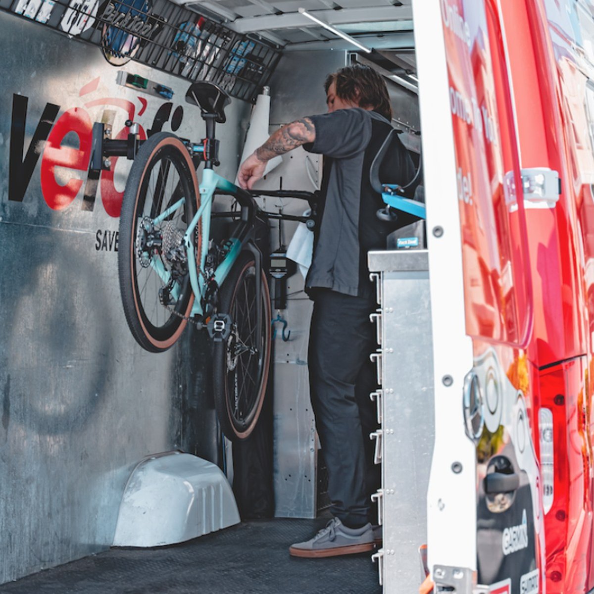 Mechanic working on a teal gravel bike mounted in a repair stand inside the open rear of a mobile service van.