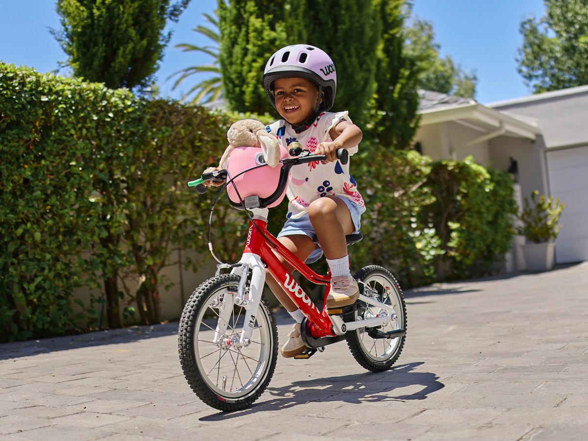 A young girl on a red woom bike with a stuffed animal in the basket attached to the handlebars.