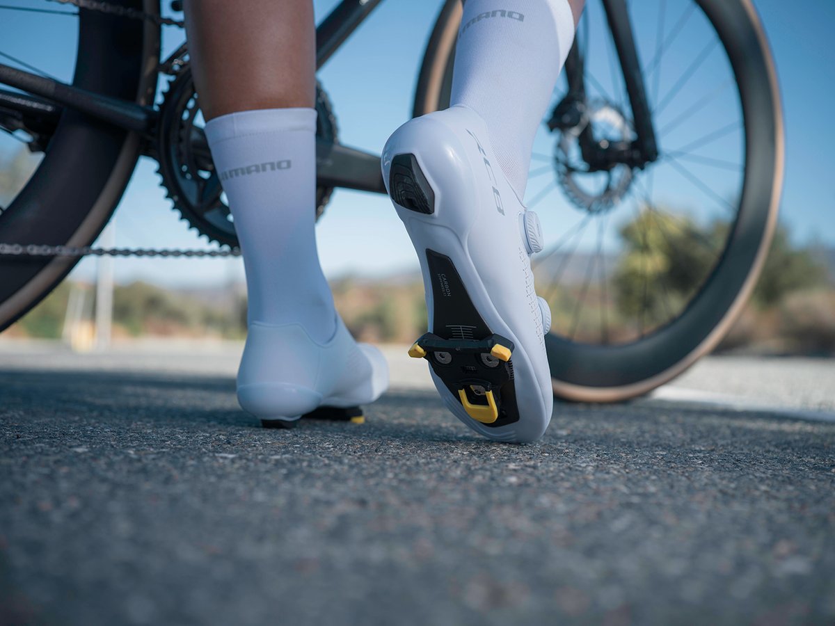 Rear view of both feet in white RC810 shoes, cleat baseplate visible on the sole unobstructed, 'SHIMANO' lettering on socks, gum-wall tire in the background