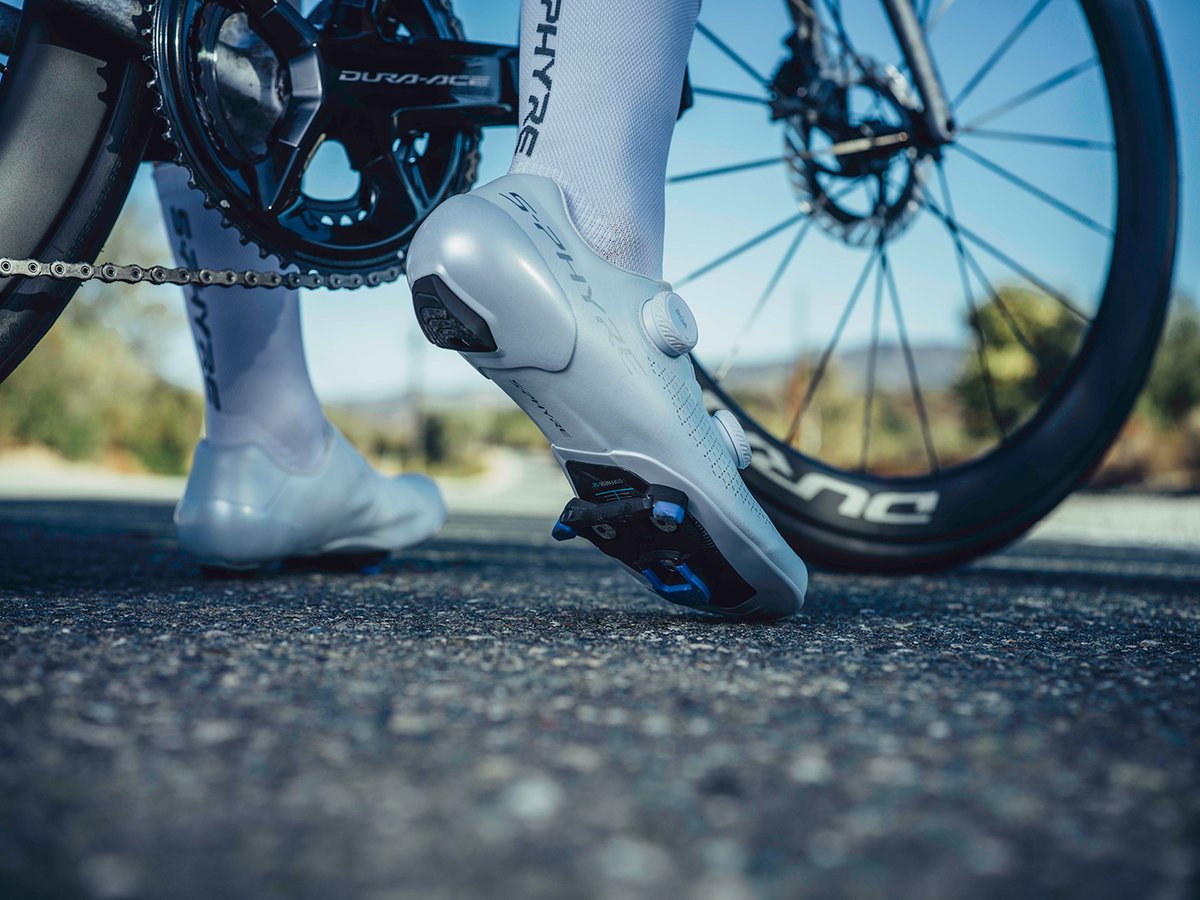 Low rear angle of a white S-PHYRE shoe clipped into a pedal, cleat baseplate visible under the sole, Dura-Ace crank and S-PHYRE sock lettering in frame