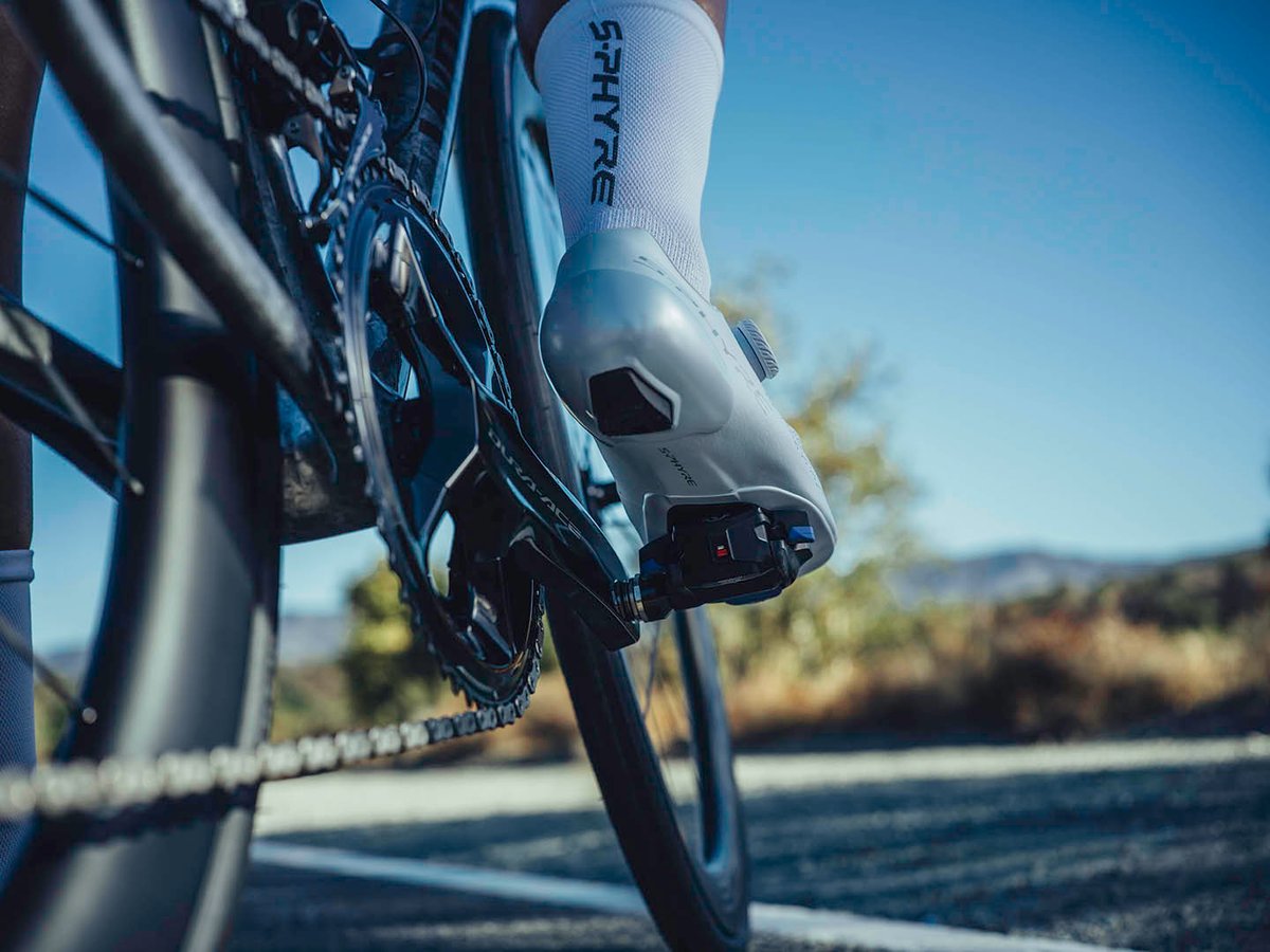 Cyclist's shoe clipped into a Dura-Ace pedal, close crop on crank and wheel, road and mountains in the background