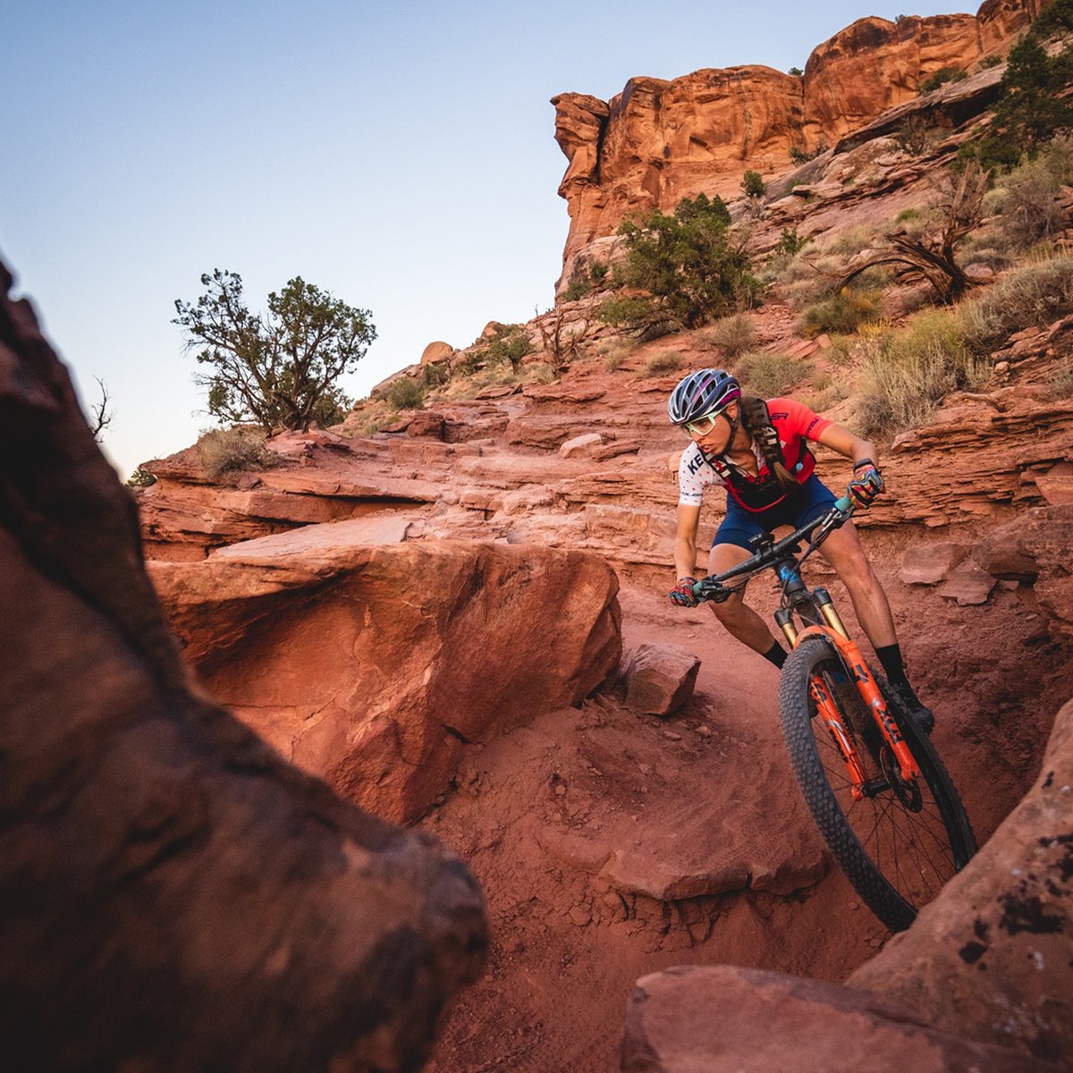 A mountain biker wearing a red and white jersey, black shorts, and a helmet rides down a steep, rocky desert trail surrounded by red sandstone formations and sparse vegetation.