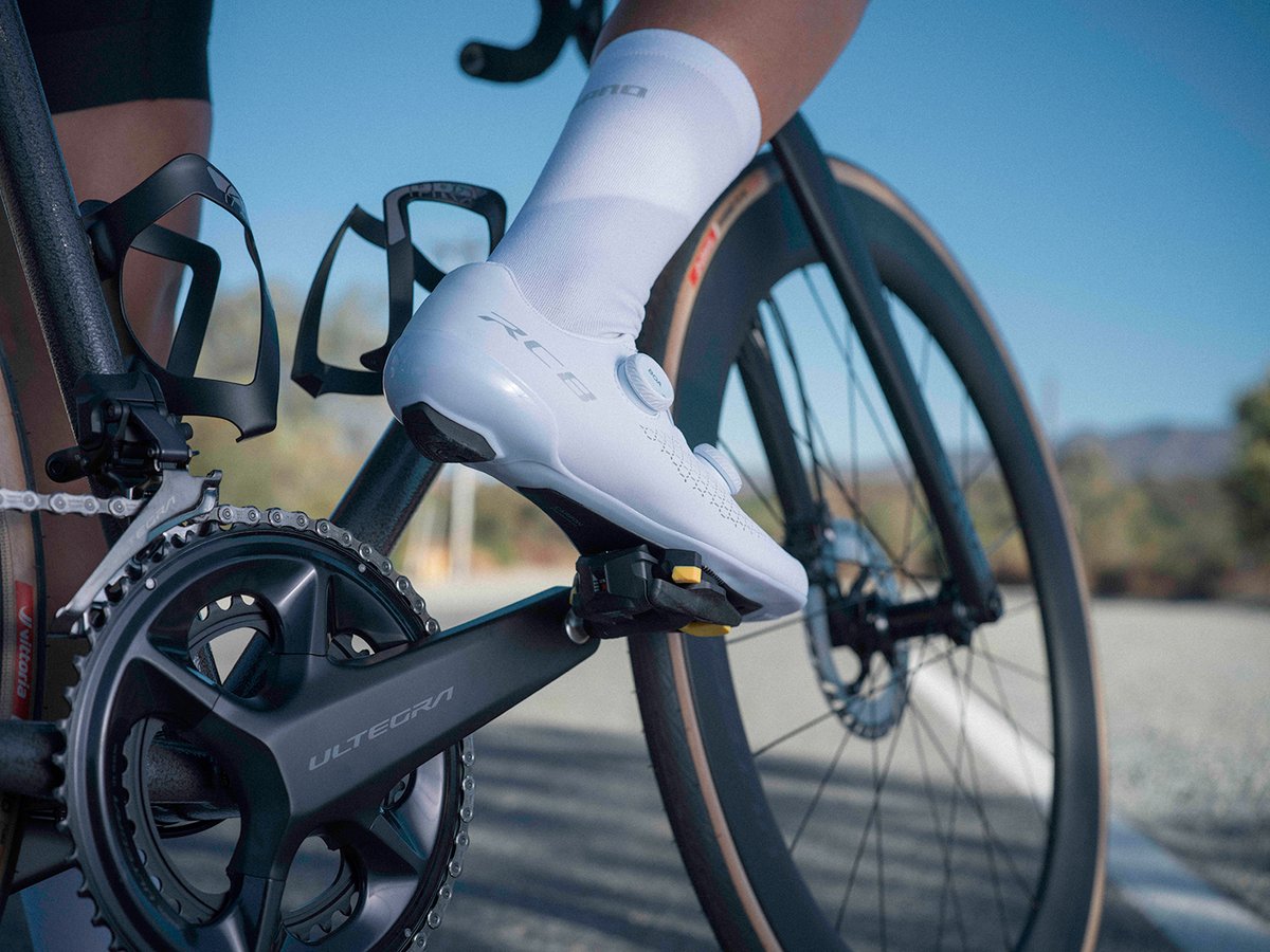 Cyclist's foot in a white RC810 shoe clipped into an Ultegra pedal, side profile showing the upper and 'RC8' lettering, wide framing with bottle cage and road bike in view