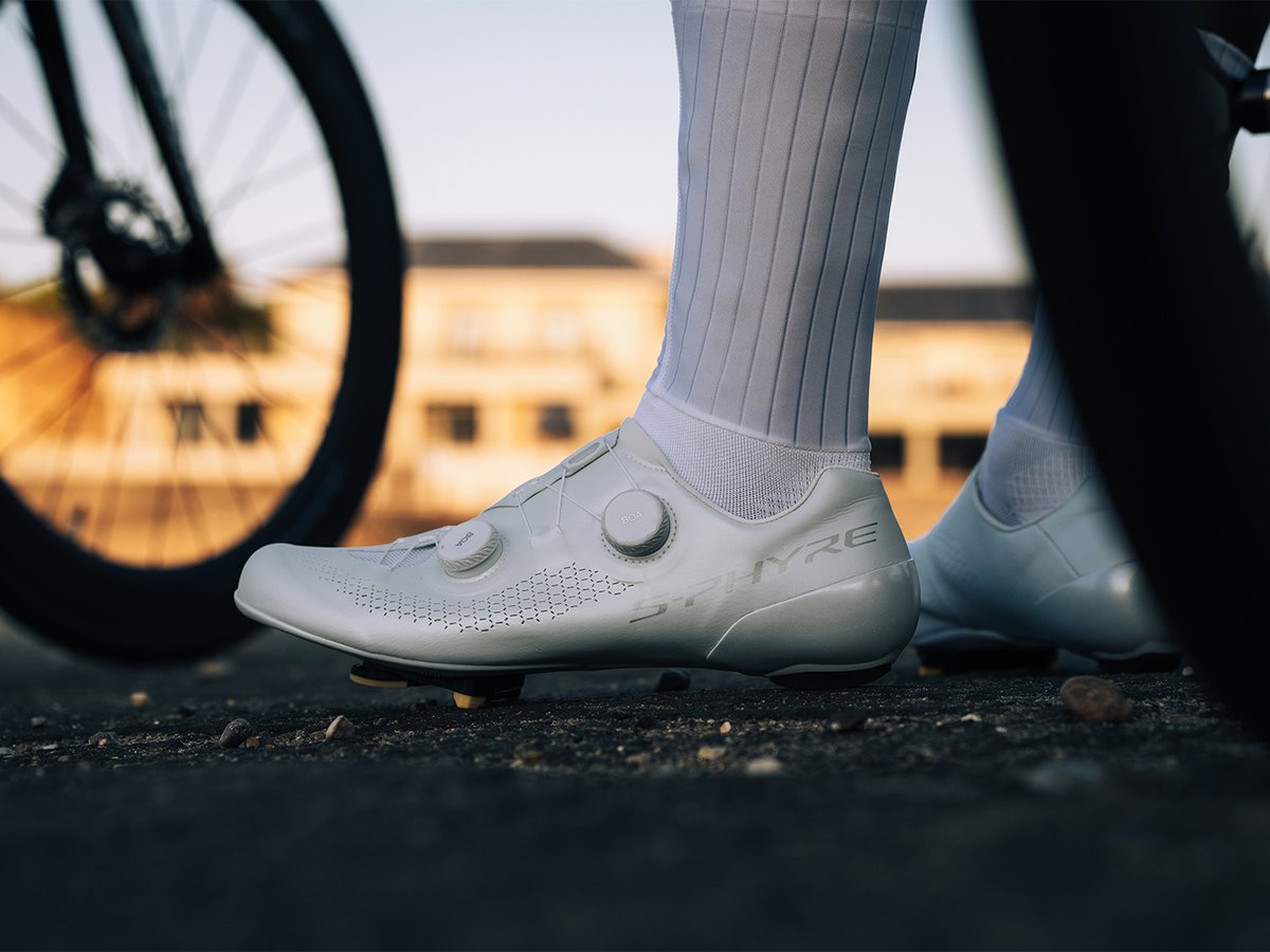 Cyclist's foot in a white S-PHYRE road shoe beside a bike wheel, cleat visible, blurred building and dusk light behind.