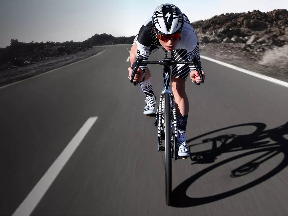 Front-on shot of a cyclist in a black and white Specialized kit riding a Tarmac SL9 toward the camera on a straight volcanic road, with shadow cast to the right.