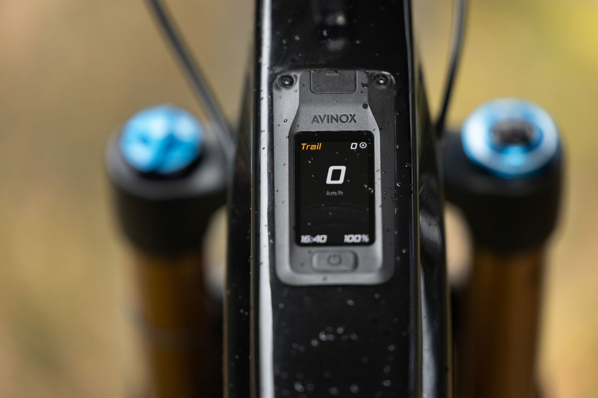 Close-up of electric mountain bike top tube display in Trail mode on rain-wet frame with blurred trail background