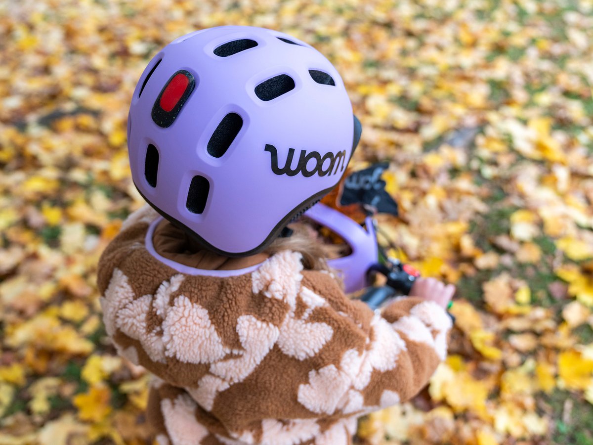 Close-up of a child wearing a lavender woom helmet, surrounded by fallen autumn leaves.