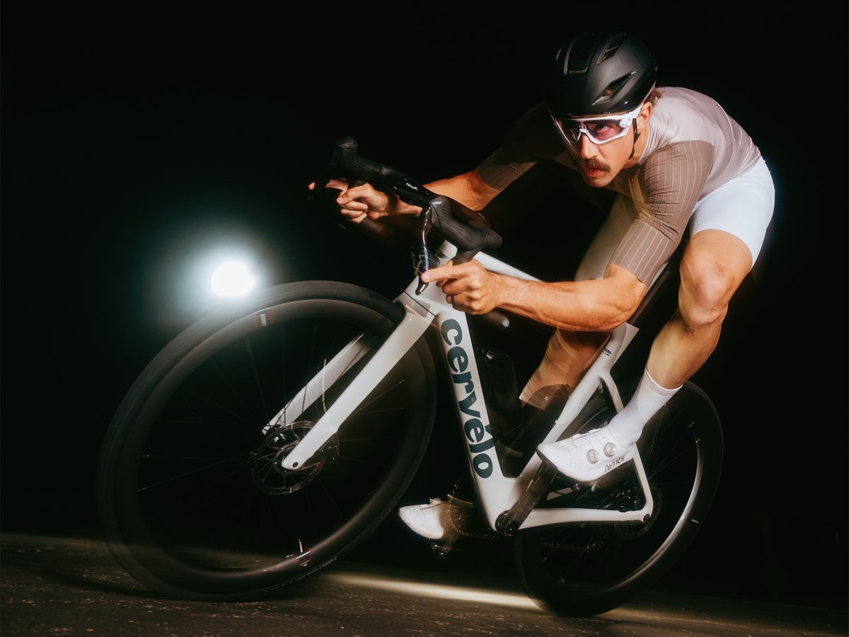 Male cyclist with a mustache in a tan Castelli kit cornering a white Cervélo road bike low against a black background, with a spotlight illuminating the front wheel.