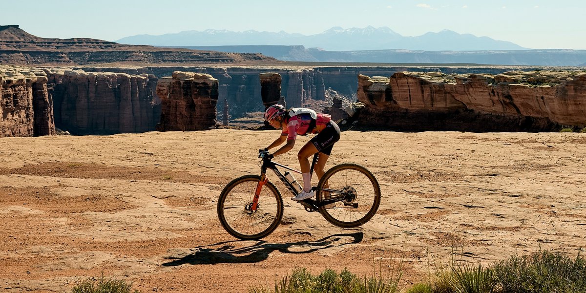Hannah Otto in a pink and black outfit rides a mountain bike across a flat, rocky desert landscape with deep canyons and distant mountains in the background.