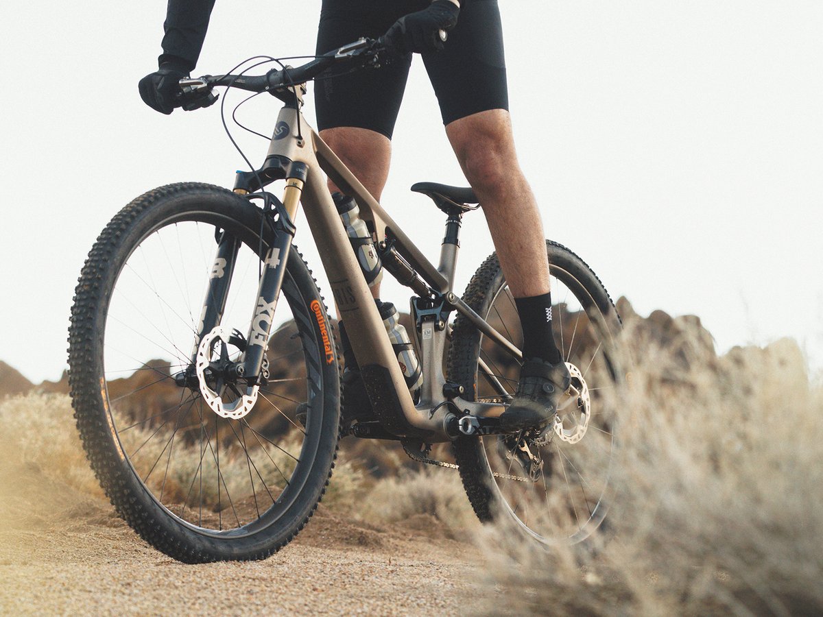 A rider wearing black cycling kit flows downhill on a matte gold Riple SL, equipped with dual water bottles and Continental tires on a dry, rocky desert trail with low shrubs and distant boulders.