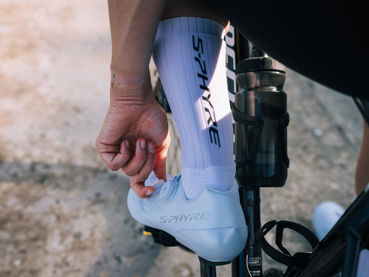 Hand adjusting a BOA dial on a white S-PHYRE shoe, S-PHYRE lettering visible on sock and shoe, water bottle in frame, warm lighting