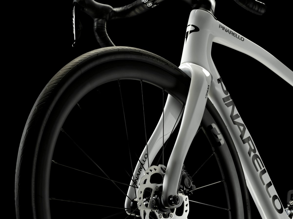 Pinarello white carbon road bike front end detail showing aero fork, disc brake rotor, drop handlebars, and deep section wheel against dark studio background