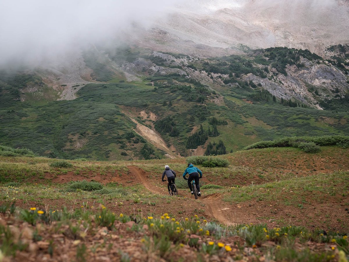 Two mountain bikers descending a red-dirt trail across an alpine meadow, low clouds hanging over the rocky peaks above.