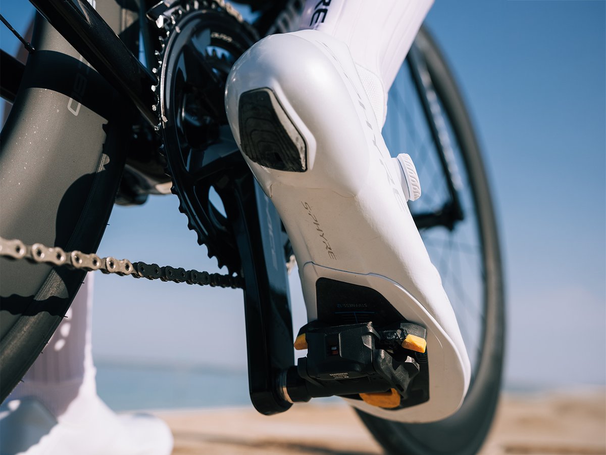 Cyclist's foot in a white S-PHYRE shoe clipped into a Dura-Ace pedal, sole profile visible against the crank, coastal water in the background