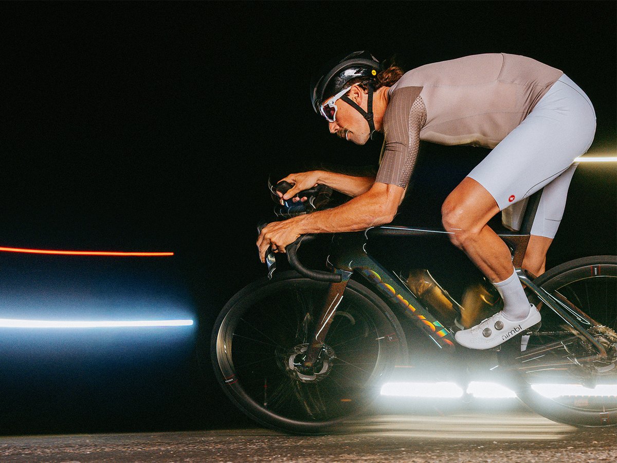 Male cyclist in a tan Castelli kit and white Nimbl shoes riding a dark Cervélo road bike in an aggressive aero position against a black background with horizontal light streaks.