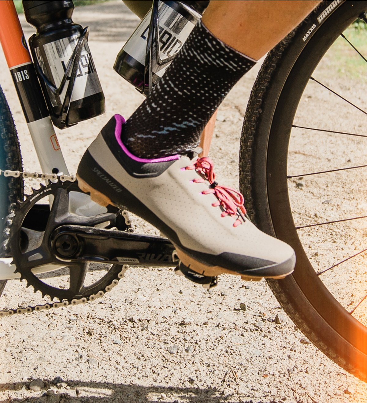 A close-up of a cyclist's foot on a bike pedal, showing a light gray cycling shoe with pink laces, black patterned socks.