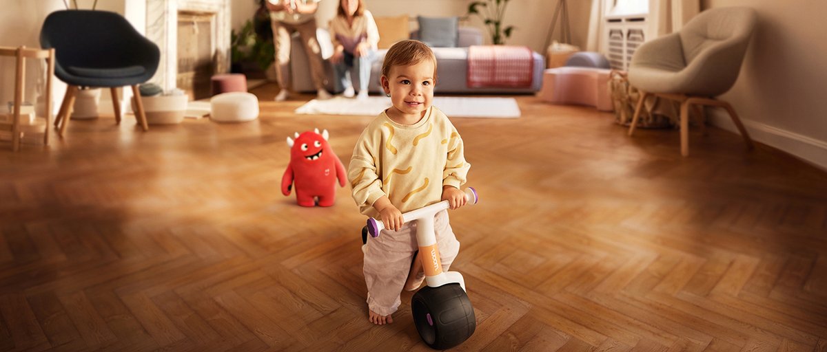 Young child riding balancing bike on their own with buddy character standing close by.