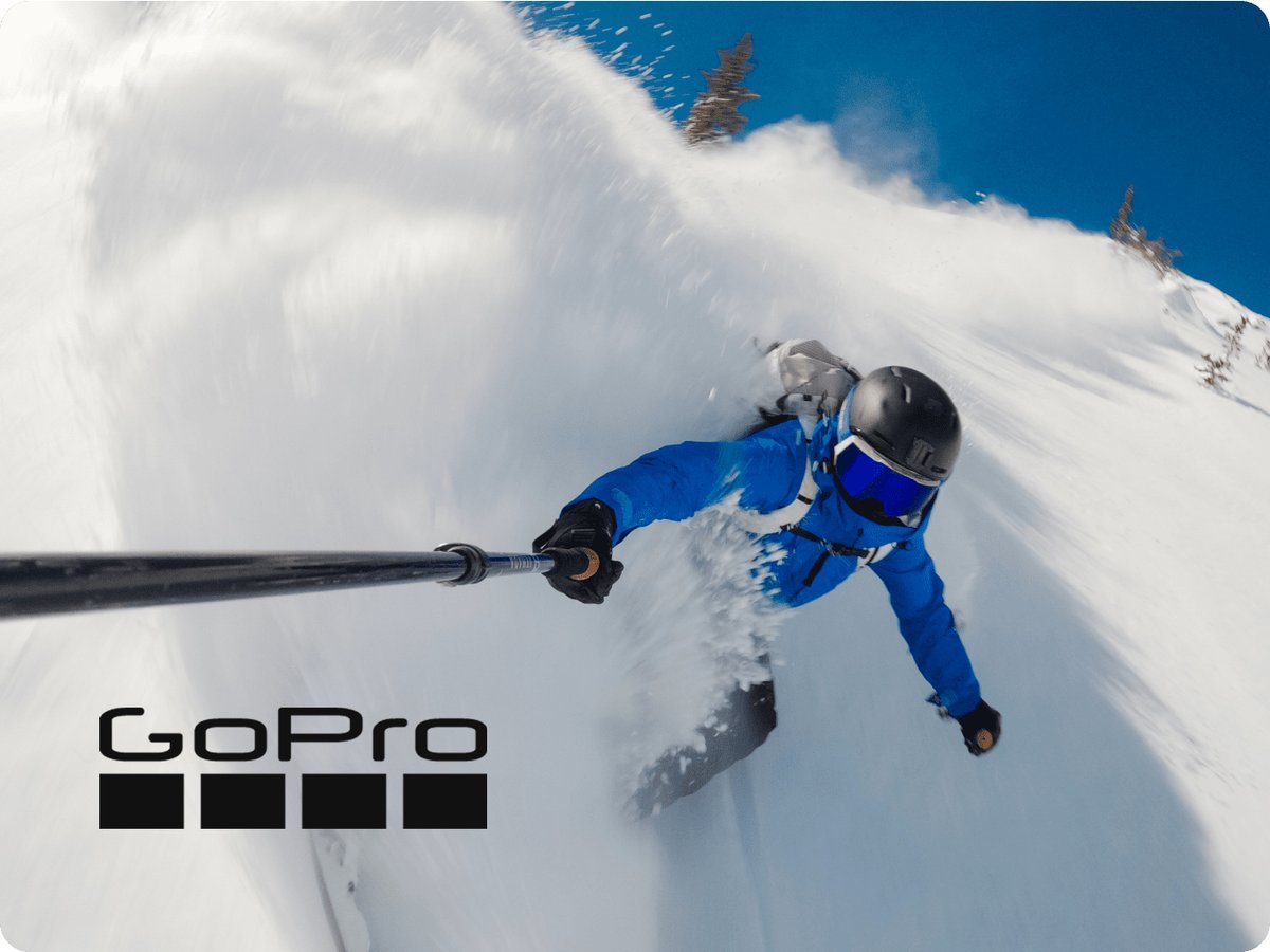 Action shot of a skier carving through deep powder while holding a pole-mounted camera, with snow spray surrounding the rider and a GoPro logo placed in the bottom-left corner.