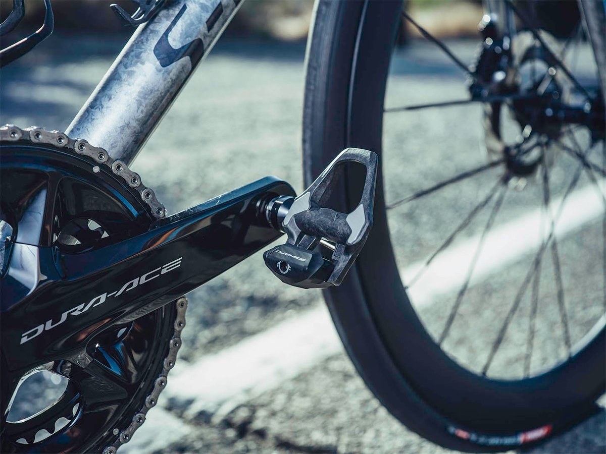 Dura-Ace PD-R9300 pedal mounted on a road bike crank, wheel and spokes visible, outdoor road setting