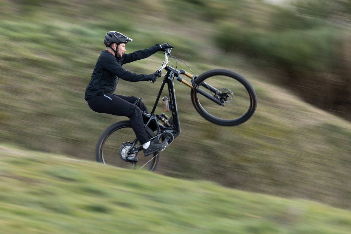 Mountain biker pulling front wheel up on electric full-suspension trail bike across grassy hillside in cool-weather riding conditions