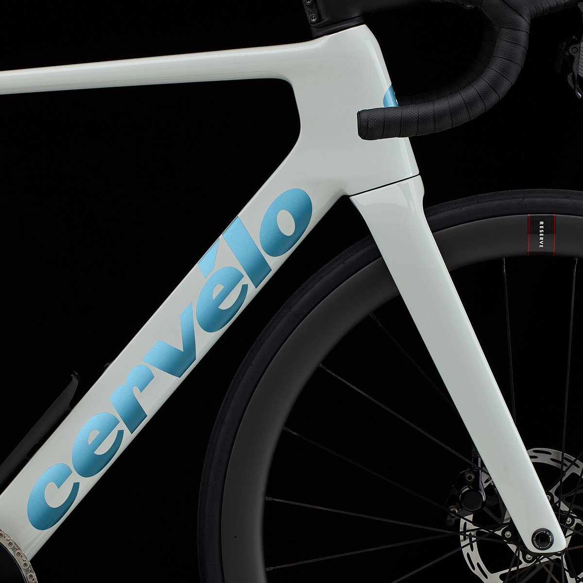 Close-up of a white Cervélo frame showing the blue "cervélo" down tube lettering, with a Reserve deep-section wheel and disc brake caliper visible behind, on a black background.