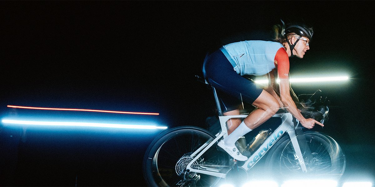 Female cyclist in an aggressive aero position on a white Cervélo Soloist bike, shot against a black background with horizontal light streaks, wearing a light blue vest and red jersey.