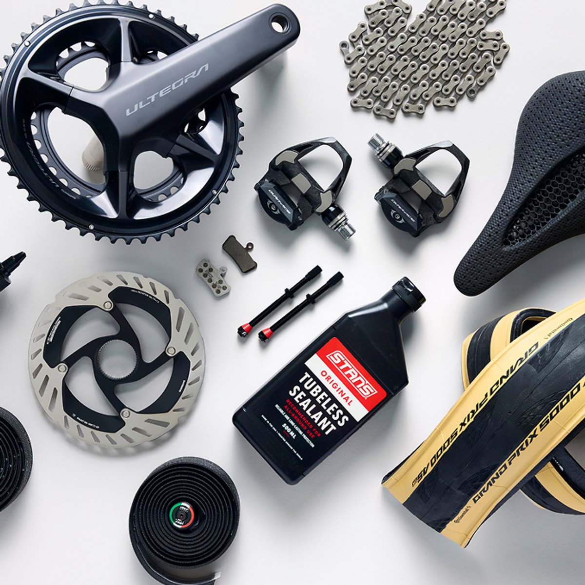 Flat-lay of road bike components — crankset, chain, cassette, pedals, saddle, brake rotor, tubeless sealant, and tire.