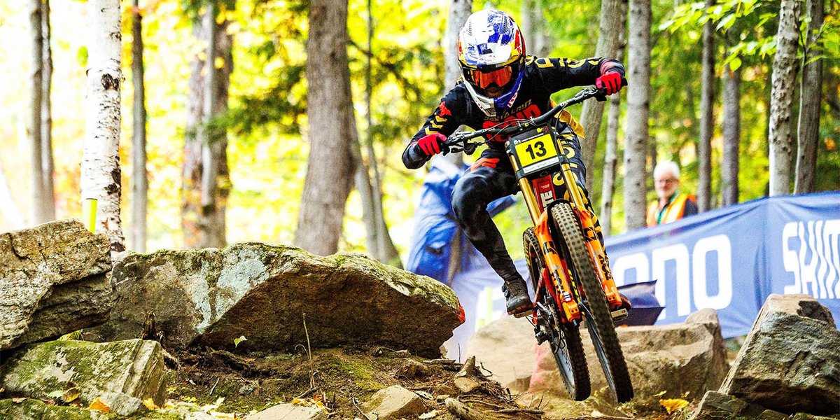 Racer on an orange and black downhill bike navigating a rocky race course through a fall foliage forest, with a course marshal visible behind.
