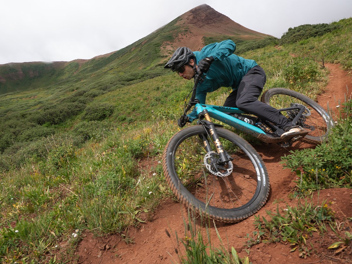 Rider carving a red-dirt switchback on a teal Yeti full-suspension mountain bike, green alpine ridgeline and low clouds behind.