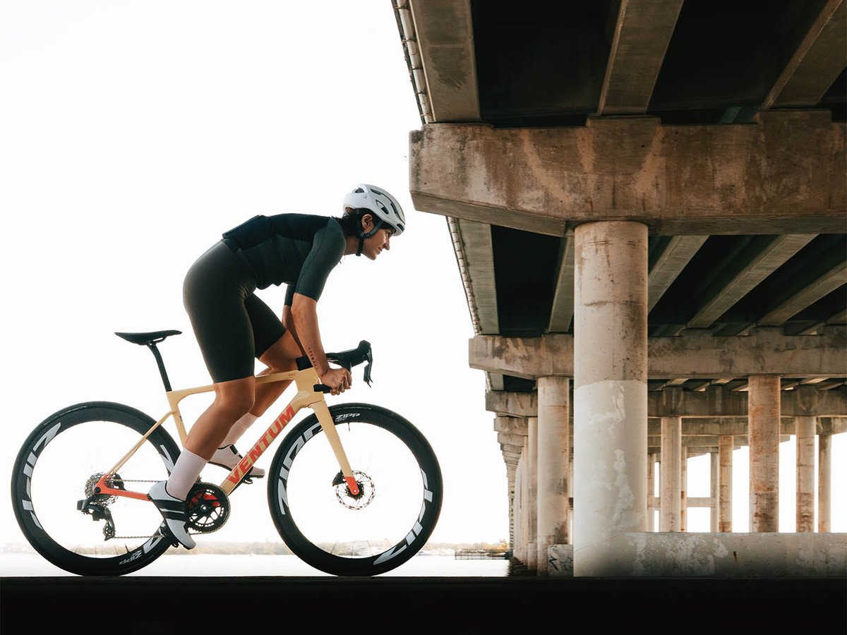 A female cyclist rides a Ventum road bike fitted with Zipp 404 wheels beneath a concrete bridge overpass, silhouetted against bright sky and open water.