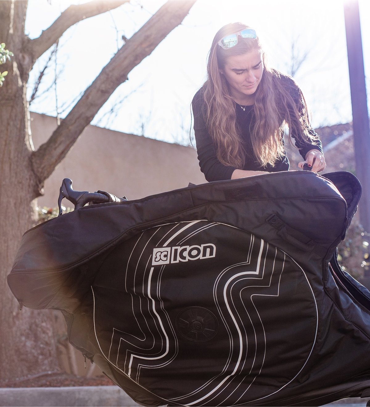 A woman with long hair and sunglasses packs her bike into a large black SCICON bike travel bag.