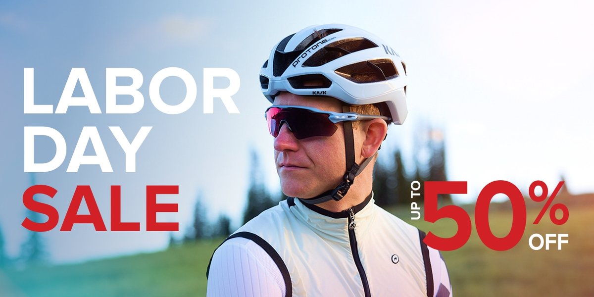 A person wearing a white and black cycling helmet, jersey, and vest stands outdoors with a blurred background of trees and sky. Text over the image reads: "Labor Day Sale. Up to 50% off.”