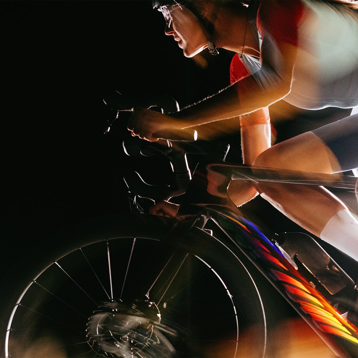 Side-profile motion shot of a female cyclist in a red and blue kit on a road bike, with multicolor light streaks across the frame against a black background.
