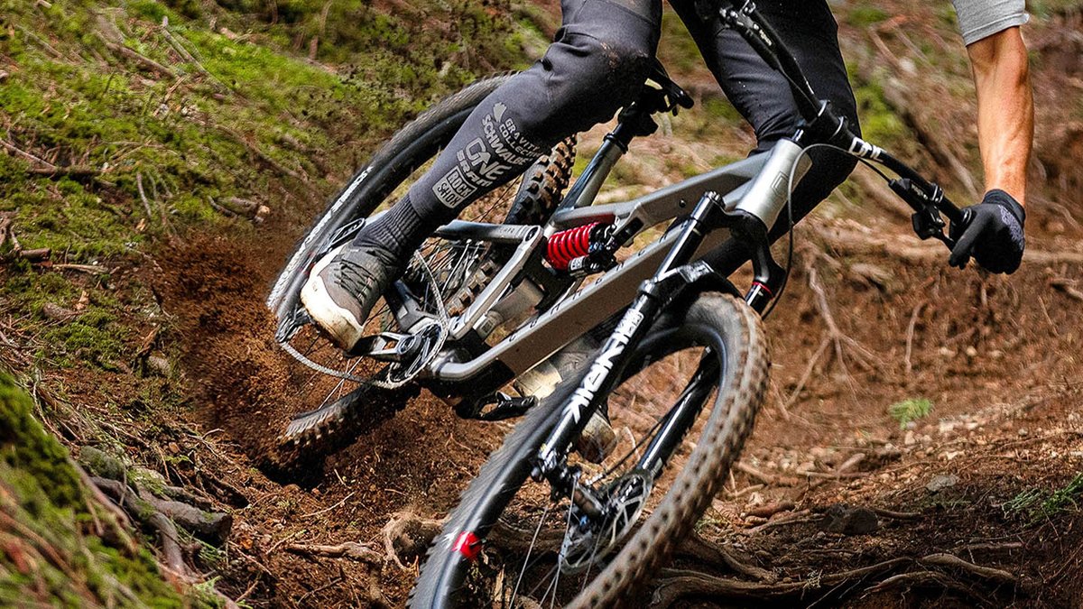 Low-angle close-up of a gray full-suspension mountain bike with coil rear shock navigating a rooted dirt berm, rear wheel digging into the soil.
