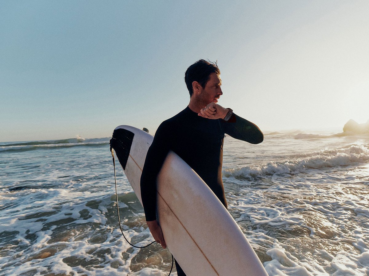 Person on beach carrying sur board talking into Garmin Fenix 9 watch.