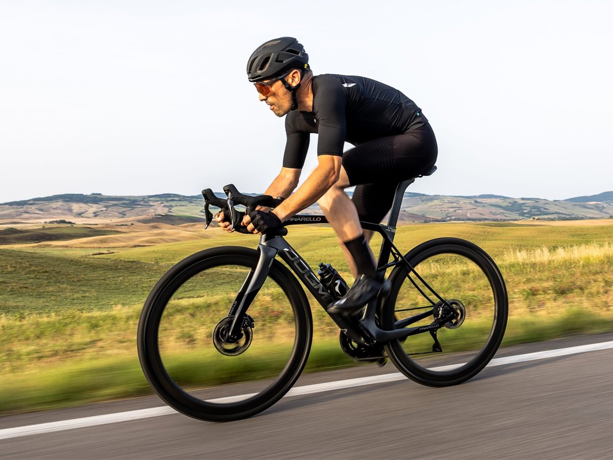 Cyclist riding Dogma road bike on paved road with hills in the background.