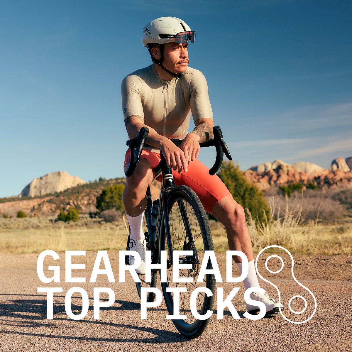 A cyclist in a tan jersey and coral bib shorts resting on a road bike on a desert highway, with red rock formations in the background. Gearhead Top Picks.