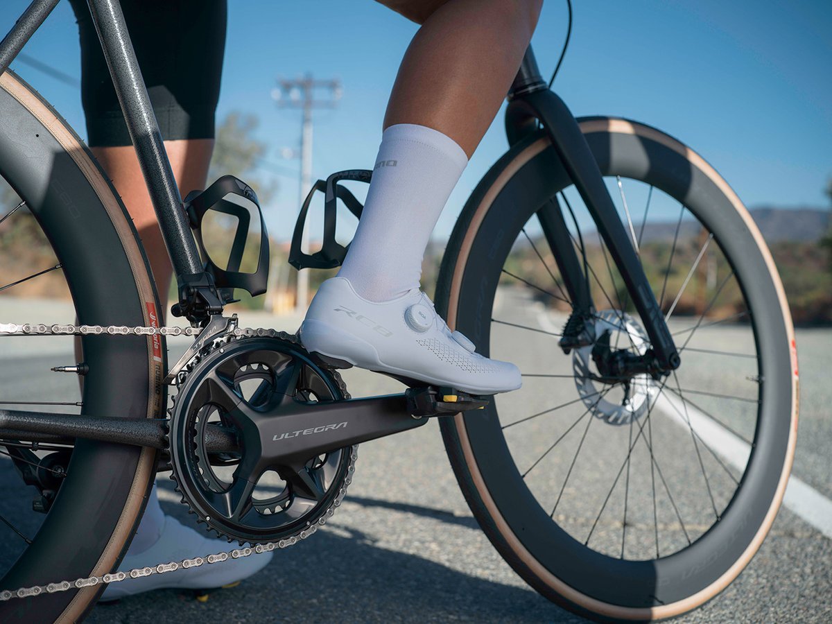 Cyclist's foot in a white RC810 shoe clipped into an Ultegra pedal, crank and cassette visible, road and hills behind.