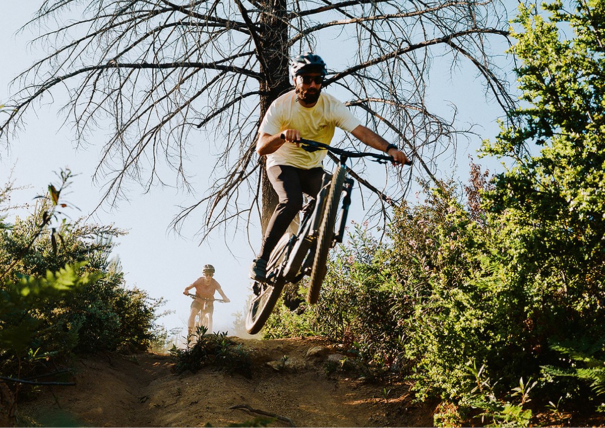 A mountain biker catches air while jumping a dirt trail, riding a full-suspension Ripmo AF. Behind, another rider wearing a helmet approaches the jump. The trail is surrounded by green bushes, and a tall, leafless tree stands in the background against a clear blue sky.