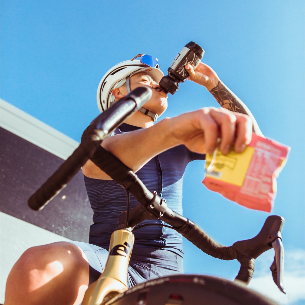 A biker in a Road Bike drinking from a bottle and holding up a ride snack package 